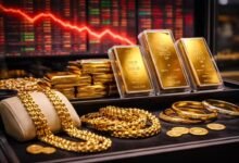 صورة سبائك ذهب ومصوغات داخل فاترينة مع خلفية شاشات بورصة حمراء تعكس هبوط أسعار الذهب عالميًا