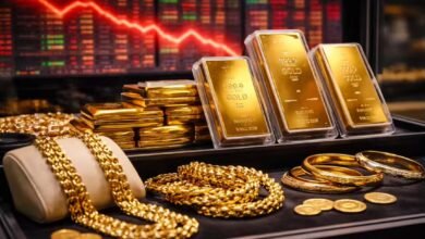 صورة سبائك ذهب ومصوغات داخل فاترينة مع خلفية شاشات بورصة حمراء تعكس هبوط أسعار الذهب عالميًا